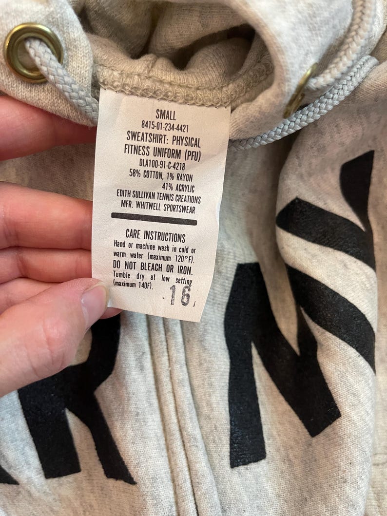 Może przedstawiać: Jasnoszara bluza z kapturem zapinana na zamek z czarnymi literami. Etykieta głosi "SWEATSHIRT: PHYSICAL FITNESS UNIFORM (PFU)" i zawiera instrukcje pielęgnacji. Bluza jest oznaczona jako "SMALL" i ma kaptur ze sznurkiem.