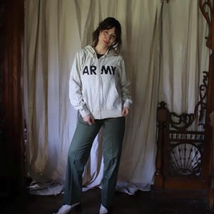 Może przedstawiać: Jasnoszara bluza z kapturem zapinana na zamek z napisem "ARMY" pogrubionymi czarnymi literami na piersi. Bluza połączona jest z oliwkowymi spodniami. Str&oacute;j jest swobodny i wygodny.