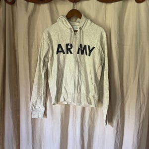 Może przedstawiać: Jasnoszara bluza z kapturem zapinana na zamek z napisem "ARMY" wydrukowanym dużymi czarnymi literami na piersi. Bluza z kapturem ma kaptur ze sznurkiem i dwie przednie kieszenie. Odzież wisi na drewnianym wieszaku.