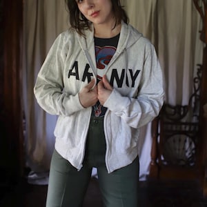 Może przedstawiać: Jasnoszara bluza z kapturem zapinana na zamek z czarnym napisem "ARMY" na piersi. Bluza z kapturem jest noszona na czarnym t-shircie z nadrukiem i połączona z oliwkowymi spodniami. Str&oacute;j jest swobodny i wygodny.