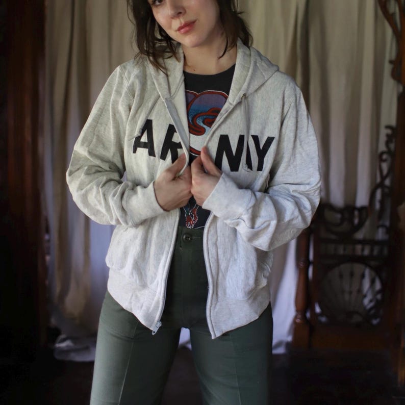 Może przedstawiać: Jasnoszara bluza z kapturem zapinana na zamek z czarnym napisem "ARMY" na piersi. Bluza z kapturem jest noszona na czarnym t-shircie z nadrukiem i połączona z oliwkowymi spodniami. Str&oacute;j jest swobodny i wygodny.