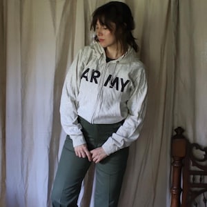 Może przedstawiać: Jasnoszara bluza z kapturem zapinana na zamek z czarnym napisem "ARMY" na piersi. Bluza posiada kaptur ze sznurkiem i przednie kieszenie. Osoba ma na sobie oliwkowe spodnie.