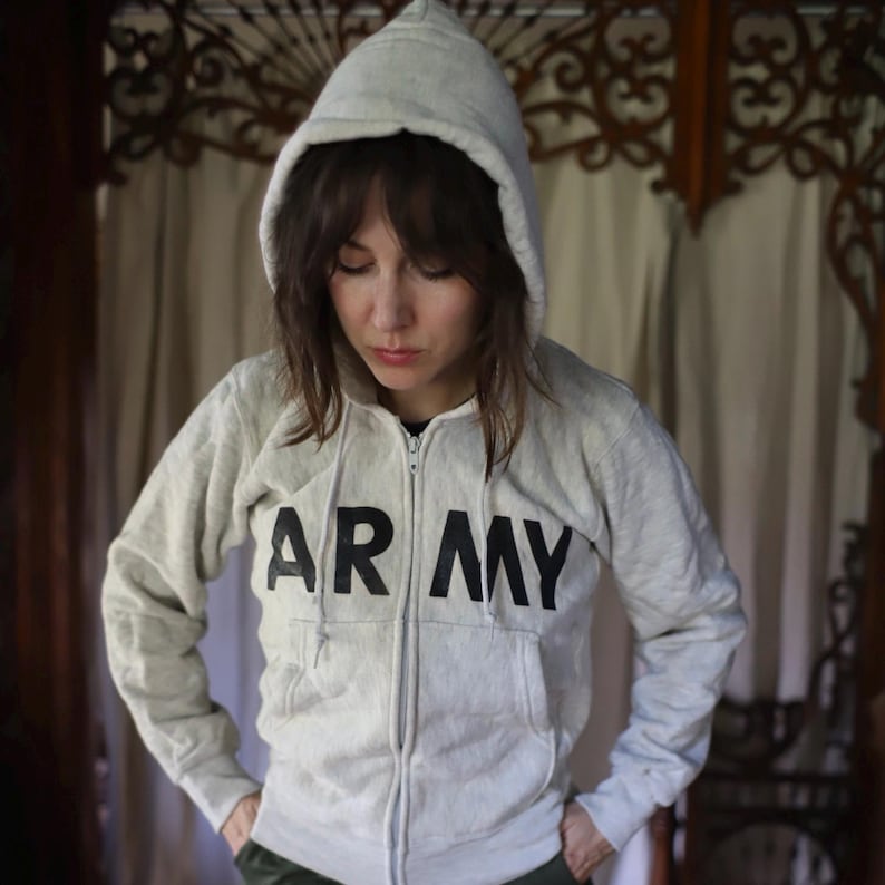 Może przedstawiać: Jasnoszara bluza z kapturem zapinana na zamek z napisem "ARMY" wydrukowanym dużymi czarnymi literami na piersi. Bluza posiada kaptur ze sznurkiem i dwie przednie kieszenie. Osoba ma na sobie bluzę.
