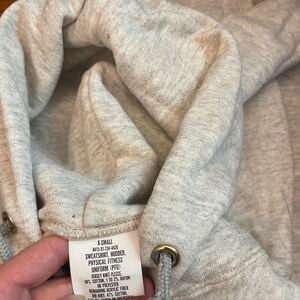 Może przedstawiać: Zbliżenie na jasnoszarą bluzę z kapturem. Kaptur ma sznurek z metalowymi oczkami. Widoczna jest metka z tekstem, w tym "SWEATSHIRT, HOODED, PHYSICAL FITNESS UNIFORM (PFU)". Tkanina wydaje się być dzianiną dżersejową.
