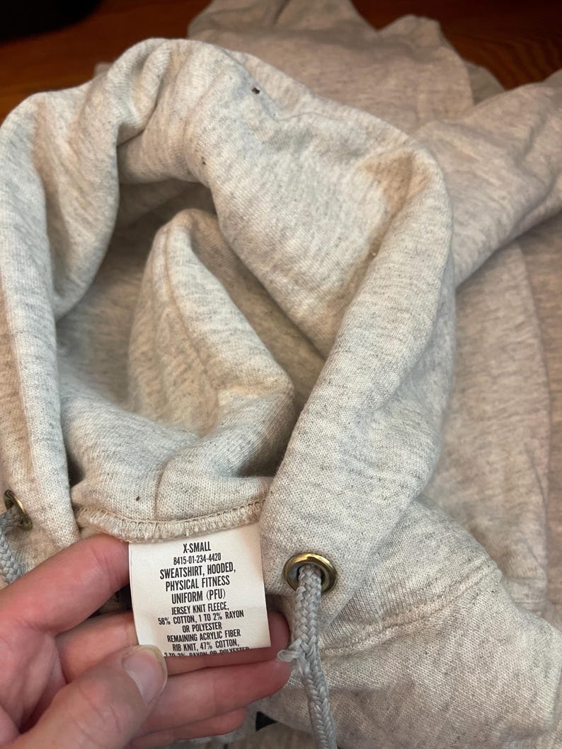 Może przedstawiać: Zbliżenie na jasnoszarą bluzę z kapturem. Kaptur ma sznurek z metalowymi oczkami. Widoczna jest metka z tekstem, w tym "SWEATSHIRT, HOODED, PHYSICAL FITNESS UNIFORM (PFU)". Tkanina wydaje się być dzianiną dżersejową.