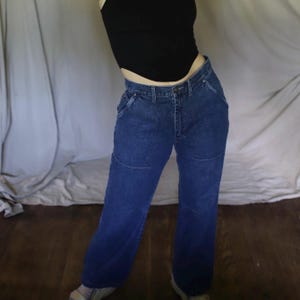 May include: Dark blue denim jeans with a high waist and a straight leg. The jeans have a classic five-pocket design and feature a unique stitched detail on the front of the legs. The denim is a medium wash, and the jeans appear to be vintage.