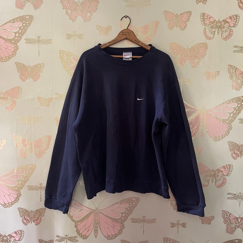 Puede incluir: Sudadera azul marino con un peque&ntilde;o logotipo blanco de Nike en el pecho. La parte superior de manga larga y cuello redondo est&aacute; colgada de una percha de madera. El fondo presenta un papel pintado con motivos de mariposas rosas y blancas.