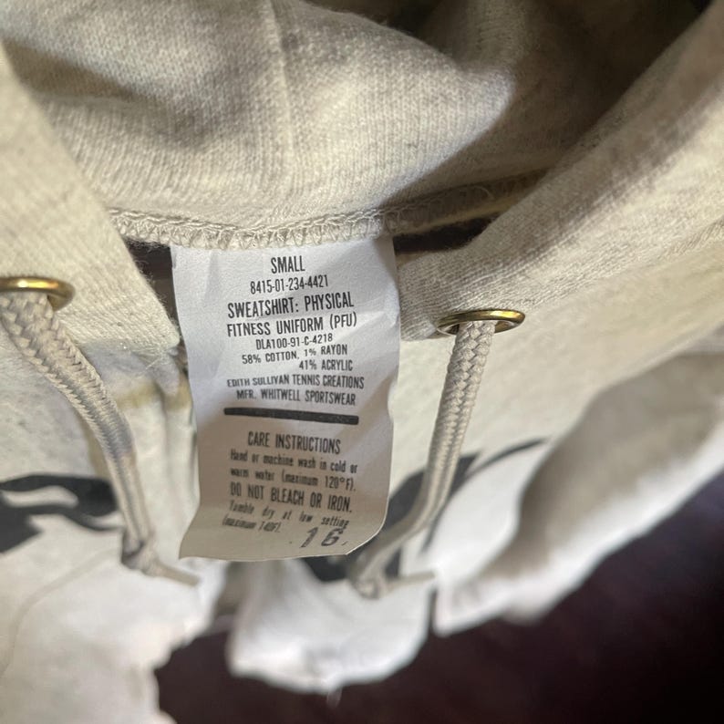 Może przedstawiać: Zbliżenie jasnoszarej bluzy z kapturem ze sznurkiem. Widoczna jest metka z napisem "SWEATSHIRT: PHYSICAL FITNESS UNIFORM (PFU)" oraz instrukcją pielęgnacji. Bluza jest oznaczona jako "SMALL".