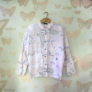 May include: A light pink button-down shirt with a map design in gray and purple. The long-sleeved shirt has a pocket on the chest and is hanging on a wooden hanger. The map design includes text such as "Atlantic Ocean" and "Caribbean Islands".