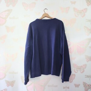 Puede incluir: Una sudadera azul marino cuelga de una percha de madera. La sudadera de cuello redondo tiene mangas largas y est&aacute; hecha de un material suave. El fondo es un papel pintado con un patr&oacute;n repetido de mariposas rosas y doradas.