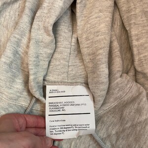 Może przedstawiać: Jasnoszara bluza z kapturem z metką z napisem "SWEATSHIRT, HOODED: PHYSICAL FITNESS UNIFORM (PFU)". Etykieta zawiera r&oacute;wnież instrukcje dotyczące pielęgnacji. Bluza jest w rozmiarze X-Small.