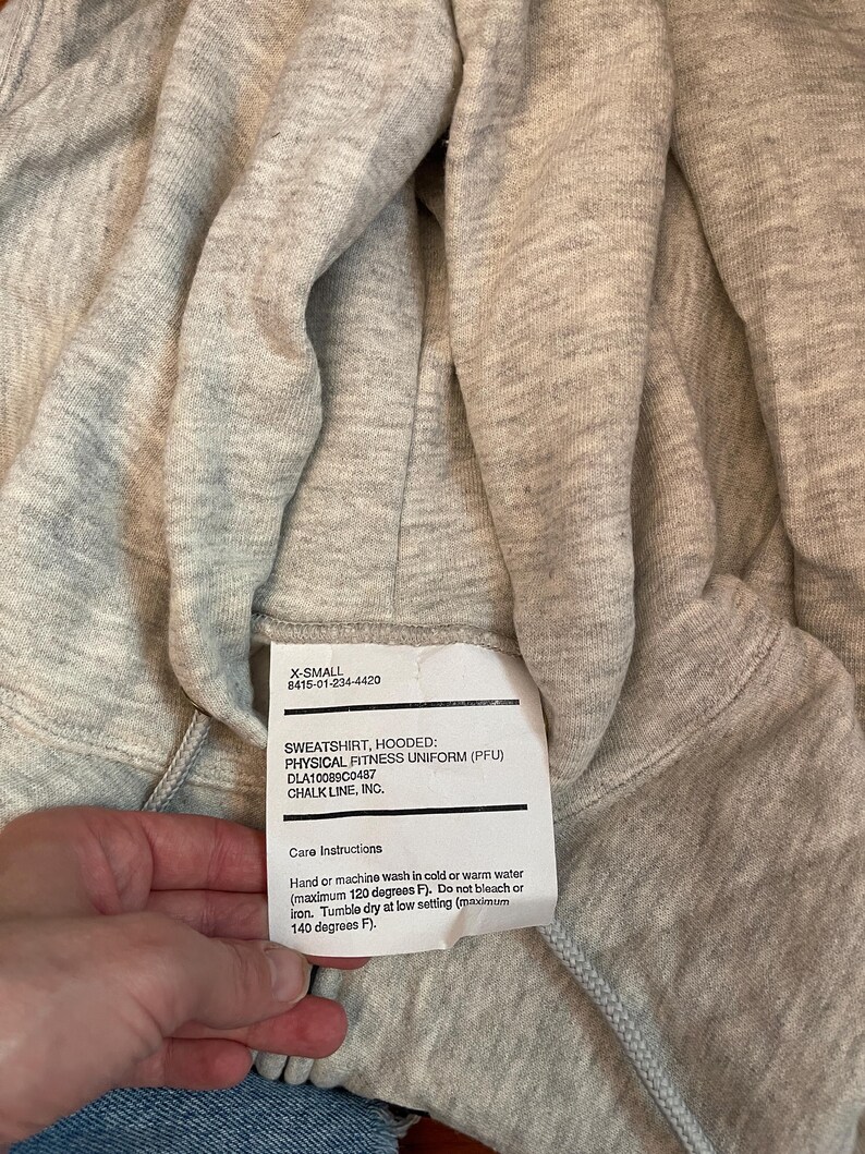 Może przedstawiać: Jasnoszara bluza z kapturem z metką z napisem "SWEATSHIRT, HOODED: PHYSICAL FITNESS UNIFORM (PFU)". Etykieta zawiera r&oacute;wnież instrukcje dotyczące pielęgnacji. Bluza jest w rozmiarze X-Small.