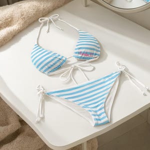 Puede incluir: Un conjunto de bikini de dos piezas con un patrón de rayas horizontales en azul claro y blanco. La parte superior triangular incluye tiras finas blancas para atar y el nombre Carlin bordado en letra rosa. La braga a juego tiene lazos laterales.