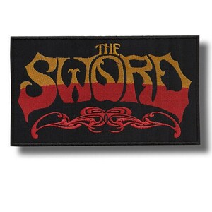May include: A rectangular black embroidered patch featuring the text THE SWORD in a stylised font. The top half of the lettering is gold, while the bottom half is red. Below the text is a symmetrical red decorative scrollwork design.