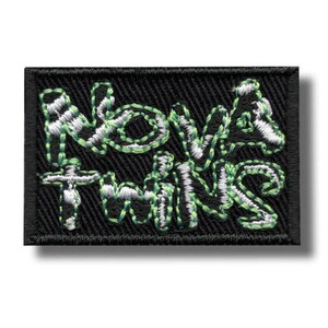 Könnte beinhalten: Ein rechteckiger, schwarz bestickter Aufnäher mit dem Text Nova Twins in einer stilisierten, von Graffiti inspirierten Schriftart. Die Buchstaben sind mit leuchtend hellgrünem Garn umrandet und weiß ausgefüllt, auf schwarzem Stoff.