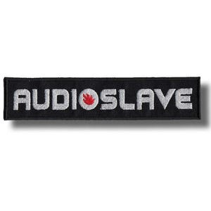 Puede incluir: Un parche rectangular bordado en negro con la palabra AUDIOSLAVE en letras mayúsculas de color gris plateado. La letra O está sustituida por un círculo blanco que contiene un pequeño icono de llama roja sobre el fondo de tela negra.