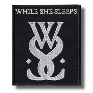 May include: A rectangular black fabric patch featuring white embroidered text that reads WHILE SHE SLEEPS at the top. Below the text is a large, stylized white embroidered logo consisting of interlocking letters and two small diamond shapes.