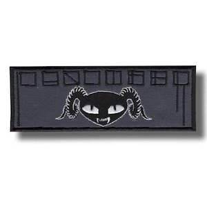 May include: A rectangular embroidered patch in dark gray with a black border. The center features a black stylized creature head with curved horns, white eyes, and sharp teeth. Above the creature are eight small black square outlines.