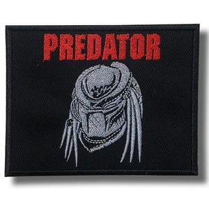 May include: A rectangular black embroidered patch featuring the word PREDATOR in bold red capital letters at the top. Below the text is a detailed grey and black embroidered graphic of the Predator creature's helmeted head and dreadlocks.