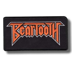 May include: A rectangular embroidered patch with a black background and a white border. The word Beartooth is stitched in a stylised orange font with a white outline, featuring sharp, angular lettering characteristic of a band logo design.