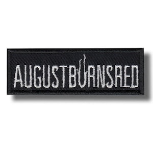 Könnte beinhalten: Ein rechteckiger, schwarz bestickter Aufnäher mit dem weißen Schriftzug AUGUST BURNS RED in einer stilisierten Schriftart. Der Aufnäher hat einen schwarz umkettelten Rand und ein flaches Design zur Anbringung auf verschiedenen Stoffen.