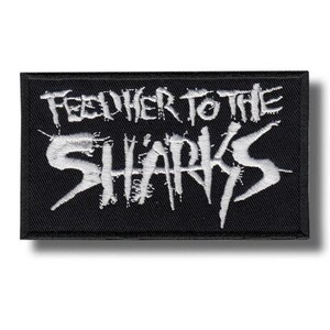 May include: A rectangular black embroidered patch featuring the white text FEED HER TO THE SHARKS in a jagged, distressed font style. The embroidery has a textured appearance with a thick black border surrounding the white lettering.