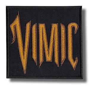 May include: A rectangular black fabric patch featuring the word VIMIC embroidered in a bold, golden-yellow thread. The lettering has a sharp, angular font style with a distinct stitched border surrounding the entire perimeter of the patch.