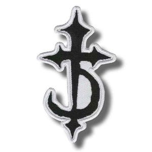 Może przedstawiać: Czarna haftowana naszywka przedstawiająca stylizowany symbol krzyża z zakrzywioną dolną częścią, obszyta białą nicią. Wzór ma ostre, spiczaste krawędzie u góry i po bokach, tworząc charakterystyczny kształt emblematu o wysokim kontraście.