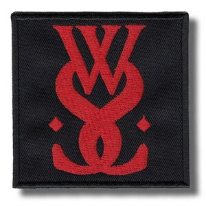May include: A square-shaped black embroidered patch featuring a bold, red stylized monogram logo in the center. The design consists of interlocking lines forming a symmetrical emblem, flanked by two small red diamond shapes on the left and right sides.