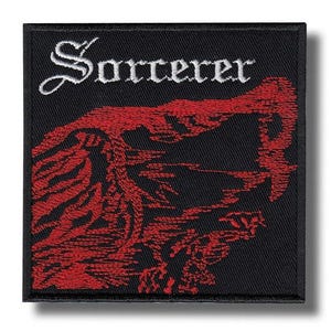 Può includere: Una toppa quadrata ricamata con sfondo nero che presenta la parola Sorcerer in lettere gotiche bianche nella parte superiore. Sotto il testo si trova un dettagliato disegno in filo rosso che raffigura un paesaggio montano scosceso.