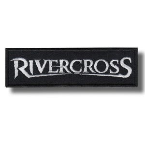 Puede incluir: Un parche rectangular bordado en negro con la palabra RIVERCROSS en letras mayúsculas blancas. El texto está estilizado con una fuente serif afilada y centrado sobre el fondo de tela negra, que tiene un borde cosido visible.