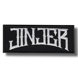 May include: A rectangular black embroidered patch featuring the word JINJER stitched in white thread. The lettering has a stylized, jagged font design with a slight arch, set against a solid black fabric background with a visible woven texture.
