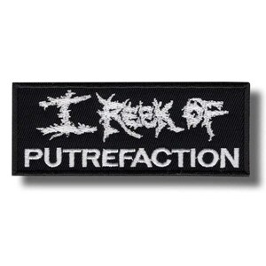 May include: A rectangular black embroidered patch featuring white thread. The top line displays the text I REEK OF in a jagged, distressed font style. The bottom line features the word PUTREFACTION in a clean, bold, sans-serif typeface.