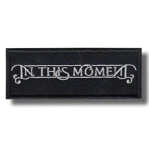 May include: A rectangular black embroidered patch featuring the white text IN THIS MOMENT in a stylised font. The lettering is enclosed within a thin white border line that extends into decorative swirls at both the left and right ends.