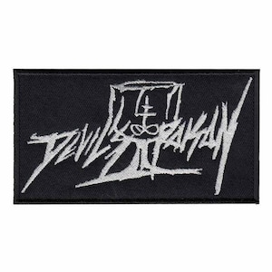 May include: A rectangular black embroidered patch featuring white thread stitching. The design displays stylized, jagged lettering that reads Devil Master, with a central symbol incorporating a cross and an infinity loop within a box frame.