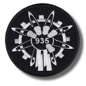 May include: A circular black embroidered patch featuring a white gear design in the background. In the center, a white circular emblem displays the number 935 surrounded by several white safety pins arranged in a star-like pattern with vertical bars.