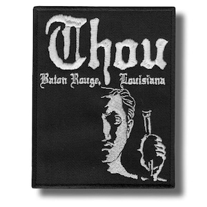 Puede incluir: Un parche rectangular negro bordado con hilo blanco. El texto Thou, Baton Rouge, Louisiana aparece en una fuente gótica. Debajo del texto hay un gráfico bordado en blanco de un rostro de perfil que sostiene un objeto con forma de botella.