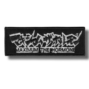 Pode incluir: Um emblema retangular preto bordado com caracteres japoneses brancos em uma fonte serrilhada e estilizada no topo, com o texto MAXIMUM THE HORMONE bordado em uma fonte branca sem serifa logo abaixo dos caracteres japoneses.