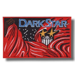 Può includere: Una toppa rettangolare ricamata con la scritta DARK STAR in lettere blu su sfondo nero. Il design include una figura avvolta in un mantello rosso con linee fluide, due stelle gialle al posto degli occhi e una bocca scheletrica bianca.