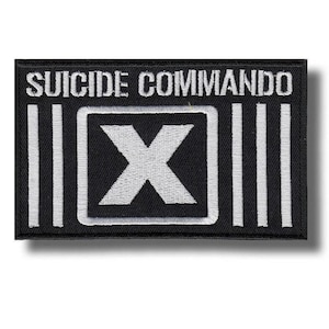 May include: A rectangular black embroidered patch featuring the white text SUICIDE COMMANDO at the top. Below the text is a white square border containing a large white letter X, flanked on both sides by three vertical white bars.
