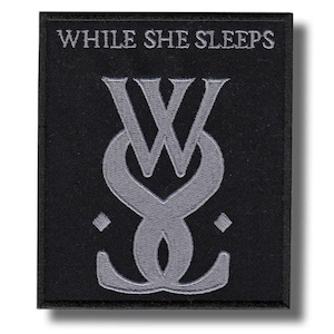 May include: A rectangular black embroidered patch featuring the text WHILE SHE SLEEPS in silver thread at the top. Below the text is a large, stylized silver monogram logo consisting of interlocking letters, flanked by two small diamond shapes.