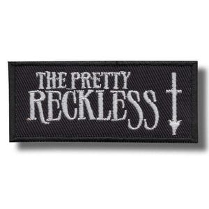 May include: A rectangular black embroidered patch featuring the white text THE PRETTY RECKLESS in a stylised serif font. To the right of the text is a vertical white symbol resembling a cross or a dagger with a pointed tip at the bottom.