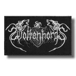 Puede incluir: Un parche rectangular negro bordado con detalles en hilo blanco. El diseño muestra un emblema central en forma de escudo flanqueado por dos cabezas de lobo estilizadas, con el texto "Wolfenhorde" escrito en una fuente gótica en el centro.