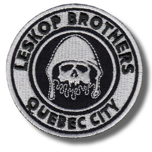 Può includere: Una toppa ricamata circolare con un design in bianco e nero. Il centro mostra un teschio con cappuccio. Il testo LESKOP BROTHERS è arcuato nella parte superiore e QUEBEC CITY nella parte inferiore, tutto in lettere nere su sfondo bianco.