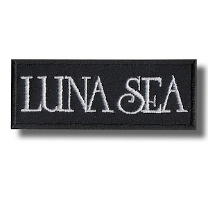 Può includere: Una toppa rettangolare in tessuto nero con il testo LUNA SEA ricamato in filo bianco. Le lettere sono in un carattere audace e stilizzato, con un bordo distinto cucito lungo tutto il perimetro della toppa per una finitura pulita.
