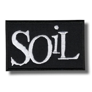 May include: A rectangular embroidered patch with a black background and white stitched lettering that reads SOIL. The letters are in a serif font, and the patch features a thick, black stitched border surrounding the entire rectangular design.