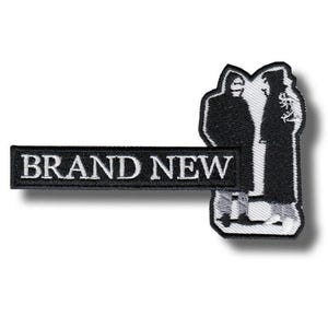 Puede incluir: Un parche bordado en blanco y negro que presenta una sección rectangular con el texto BRAND NEW en letras mayúsculas blancas, unido a una forma vertical que representa dos figuras silueteadas con abrigos largos sobre un fondo claro.