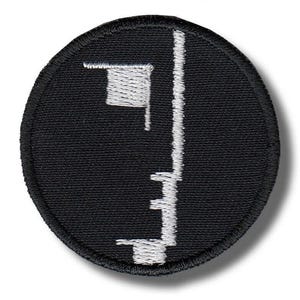 May include: A circular embroidered patch featuring a black background with a white stitched geometric design. The design consists of a small square shape in the upper left quadrant and a vertical line with stepped horizontal segments extending downward.