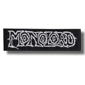 Puede incluir: Un parche rectangular bordado en negro que presenta la palabra MONOLORD en una fuente blanca estilizada y dentada. Las letras tienen una apariencia gruesa y texturizada con un contorno distintivo sobre un fondo de tela negra con bordes cosidos.