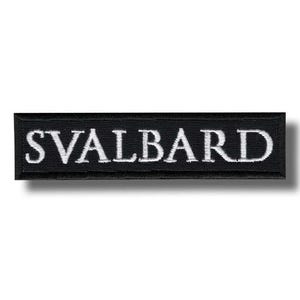 May include: A rectangular embroidered patch with a black background and a white border. The word SVALBARD is stitched in white capital letters across the center of the patch. The design features a clean, bold font with a textured fabric finish.